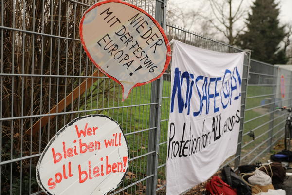 Zwei Schilder mit &quot;Nieder mit der Festung Europa!&quot; und &quot;Wer bleiben will soll bleiben!&quot; sind übereinander in einen Zaun gesteckt. Daneben hängt ein Transparent: &quot;Not safe @ALL - Protection for ALL refugees&quot;