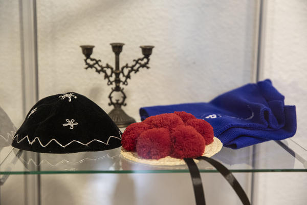 Eine Vitrine mit Kerzenleuchter hinten, blaues Tuch rechts, vorne links eine schwarze Kipa, mittig vorne eine rote Bollenhut-Kipa.