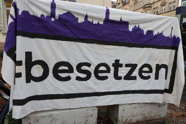 Ein Banner, auf dem &quot;#besetzen&quot; steht und eine Stadtsilhouette in violett gemalen ist.