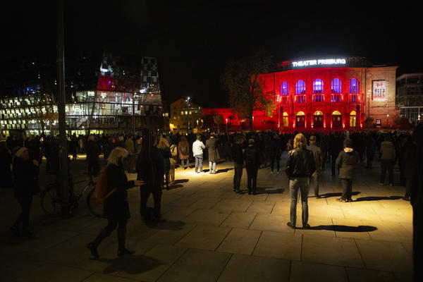 Das Freiburger Stadttheater rot beleuchtet, im Dunkeln, Auf dem Platz davor verteilt stehen Menschen mit Instrumenten.