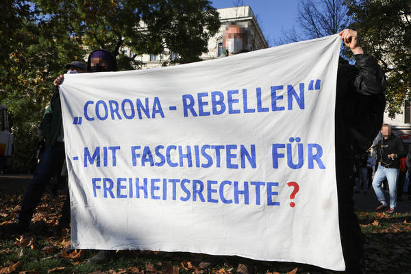 Ein weißes Transparent wird gehalten, auf dem mit blauer Schrift steht: &quot;Corona-Rebellen - Mit Faschisten für Freiheitsrechte?&quot;