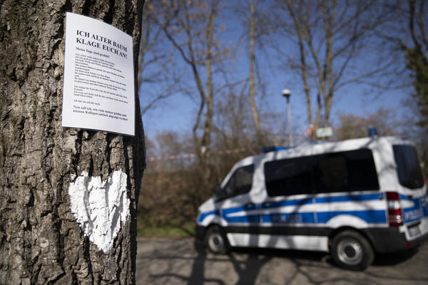 Im Vordergrund: Ein Baumstamm, an dem ein weißes Herz gemalt ist und ein Bild mit einem Gedicht hängt. Im Hintergrund eine Polizei-Wanne und ein mit Flatterband abgesperrter Bereich.