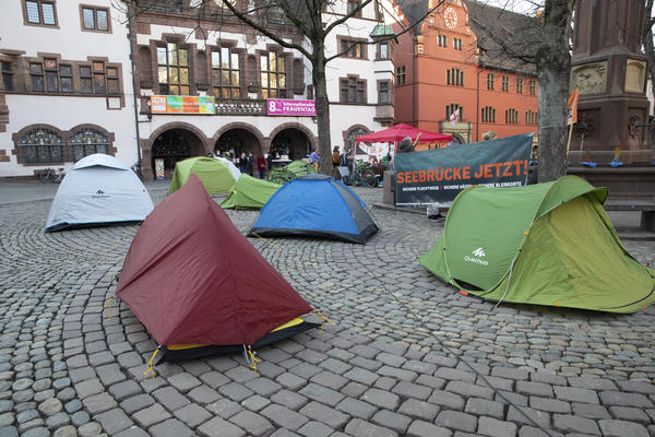 Ein paar Zelte stehen vor einem &quot;Seebrücke jetzt!&quot;-Transparent auf dem Freiburger Rathausplatz. Im Hintergrund sieht man Menschen, einen Pavillion und das Alte Rathausgebäude.