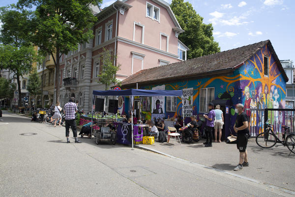 Vor dem kleinen, bunten Haus der Gartenstraße19 steht ein blauer Pavillon und Menschen stehen herum. oder sitzen. Eine Person spricht in ein Mikrofon.