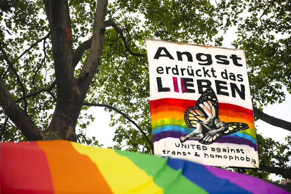 Hinter einem regenbogenfarbenen Pavillondach hängt ein senkrechtes Transparent in den Bäumen auf dem steht: Angst erdrückt das Lieben - united against trans* &amp; homophobia.