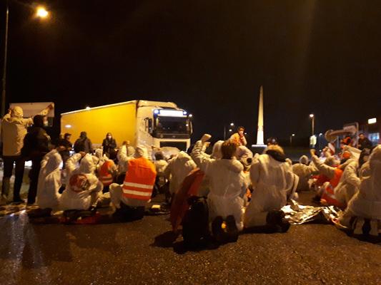 Eine Gruppe von Aktivist:innen sitzt und blockiert in weißen Malereianzügen in der Nacht, vor einem LKW.