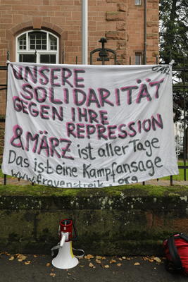 Ein Transparent hängt an einem Geländer, darunter steht ein rot-weißes Megafon. Auf dem Transparent steht: Unsere Solidarität gegen ihre Repression - 8. März ist aller Tage. Das ist eine Kampfansage (www.frauenstreik.org)
