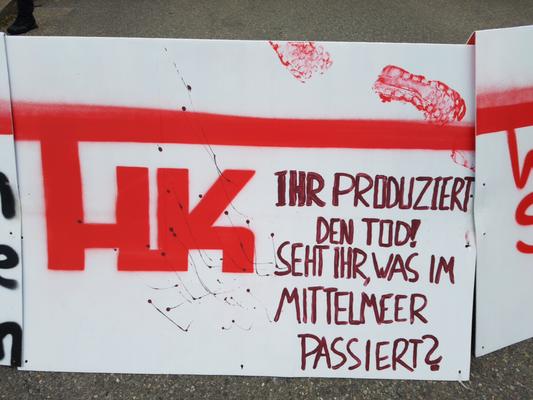 Ein weißes Schild mit symbolischen Blutflecken &amp; mit roter Farbe steht geschrieben: &quot;HK - Ihr produziert den Tod! Seht ihr, was im Mittelmeer passiert?&quot;