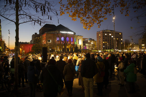 Überblicksfoto von der Gedenkkundgebung auf dem Platz der Alten Synagoge in Freiburg. Im Dunkeln stehen mehrere Hundert auf dem Platz. Im Hintergrund ist das Stadttheater zu erkennen. &quot;Wir gedenken den Opfern der Shoa&quot; steht auf einem Plakat.