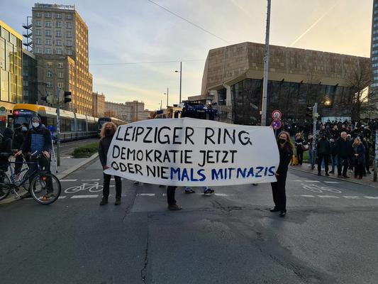 Auf dem Leipziger Ring stehen Antifaschist:innen mit einem weißen Transparent, auf dem steht: &quot;Leipziger Ring - Demokratie jetzt oder nie|mals mit Nazis.&quot; Das &quot;mals mit Nazis&quot; ist in blauer Schrift hervorgehoben.