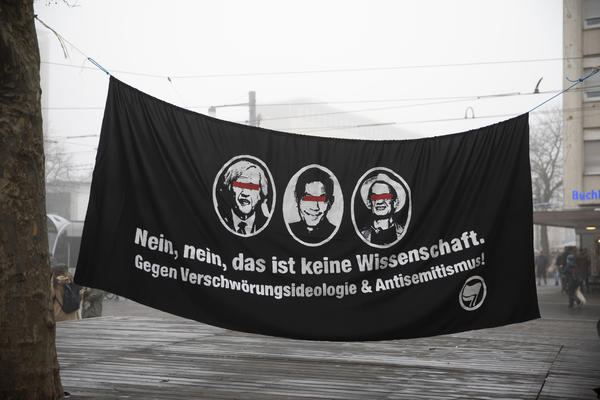 Schwarzes Transparent aufgehängt. Auf dem sind Portraits von Wodarg, Bhakdi und Schiffmann in ovalen Kreisen abgebildet. Darunter steht: &quot;Nein, nein, das ist keine Wissenschaft. Gegen Verschwörungsideologie &amp; Antisemitismus!&quot;