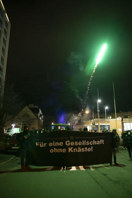 Demonstration vor der Freiburger JVA. Schwarzes Transparent mit weißer Schrift: Für eine Gesellschaft ohne Knäste! Ein grünes Feuerwerk steigt in den Himmel und beleuchtet die Umgebung grün. Im Hintergrund: mit Strahlern beleuchteter Eingang zur JVA.