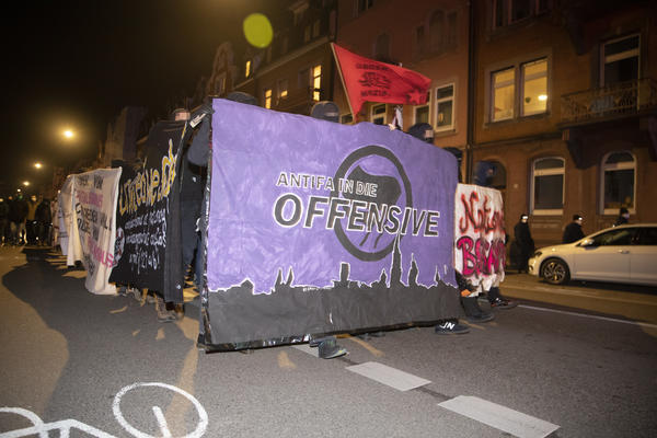 Demofront der Antifademo. Lilanes Transparent mit schwarzem Antifalogo und Stadt-Sillouette. &quot;Antifa in die Offensive&quot; steht darauf. Eine rote Fahne auf der &quot;Gegen Nazis&quot; steht wird dahinter geschwenkt.