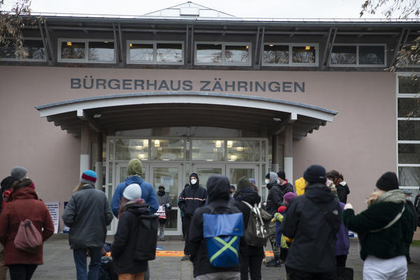 Blick auf&#039;s Bürgerhaus Zähringen. Leicht rosa Fassade. Bogenförmige Überdachung des Eingangs. Im Vordergrund eine Menschenmenge.