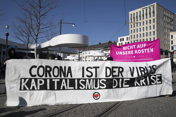 Transparent auf dem Europaplatz. Auf weißem Stoff steht &quot;Corona ist der Virus - Kapitalismus ist die Krise&quot;. Auf der Unterseite ist mittig ein Antifa-Logo angeordnet. Hinder dem Transparent sieht man ein pinkes Hochtransparent, mit dem Demomotto.