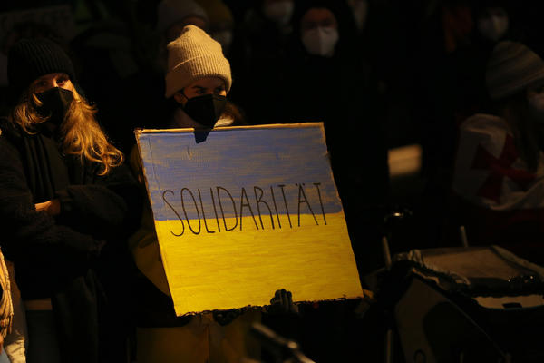 Eine Demonstrantin hält ein Schild in der Hand, das in den Farben Ukraines bemalt ist (blau-gelb). In der Mitte des Schildes steht &quot;Solidarität&quot;. Die Person stehtleicht links versetzt im Zentrum der Nahaufnahme. Es ist dunkel.