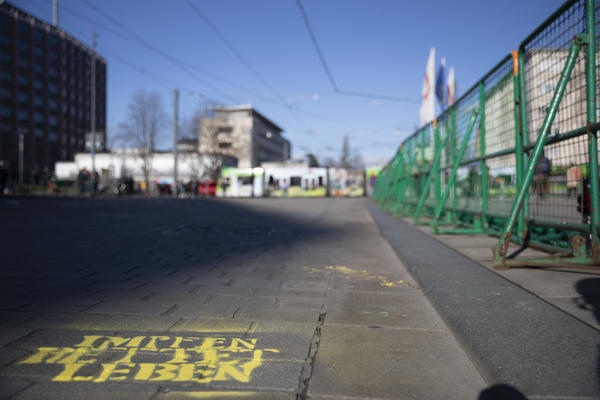Linksunten im Bild: &quot;Impfen rettet Leben&quot; mit gelber Sptrühfarbe als Stencil auf den Boden des Rotteckrings vor dem Platz der Alten Synagoge. Rechts Hamburger Gitter.