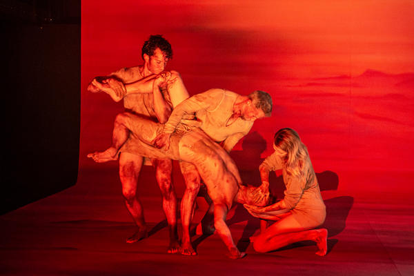 Schauspieler*innen auf der Bühne halbnackt, schmutzig und in rotes Licht getaucht vollführen zu viert eine groteske Figur