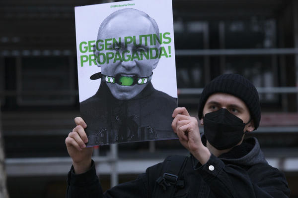 Ein männlich gelesener Demonstrant mit schwarzer Mütze wie Maske hält ein Schild in die Höhe, auf dem Putin mit einem Knebel abgebildet ist. Es steht &quot;Gegen Putins Propaganda!&quot; darauf.