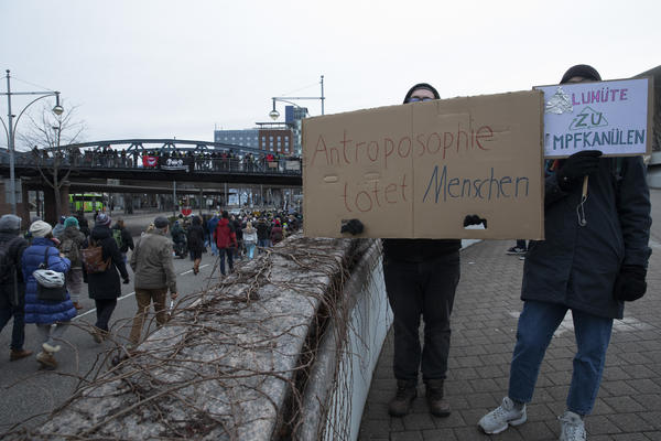 Zwei Antifaschist*innen stehen auf der Auffahrt zur Blauen Brücke. Sie tragen Schilder, 1 gegen Anthroposophie und 1 für das Impfen. Links läuft eine Querdenken-Demo vorbei.