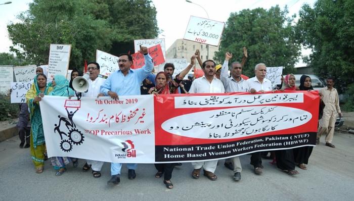 Protestmarsch mit vielen Männern und Frauen in Karatschi 2019. Sie tragen vor sich ein Banner mit den Logos der Gewerkschaftsvereinigungen