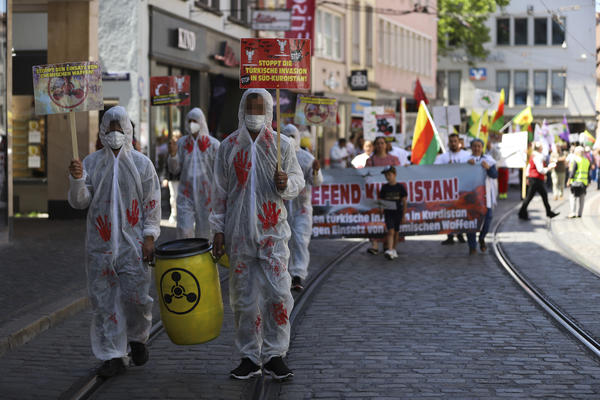 Im Vordergrund und der linken Bildhälfte laufen zwei Personen mit rot beschmierten Malanzügen und ein gelbes Fass mit schwarzem, runden Symbol darauf, vor einem Demonstrationszug, den man verschwommen im Hingergrund erkennt.