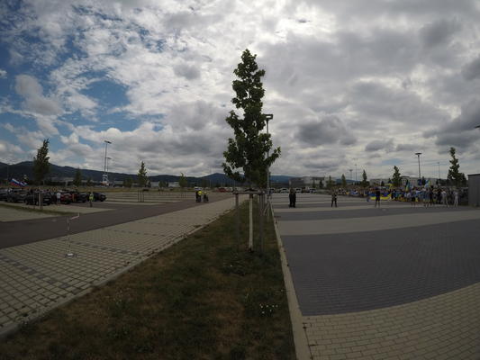 Weitwinkelaufnahme eines Parkplatzes, der druch einen Grünstreifen mit Baum in der Mitte geteilt ist. Am linken Bildrand sind Autos mit Russland-Flaggen zu sehen, auf dem rechten Bildrand Menschen mit Ukraine-Flaggen.
