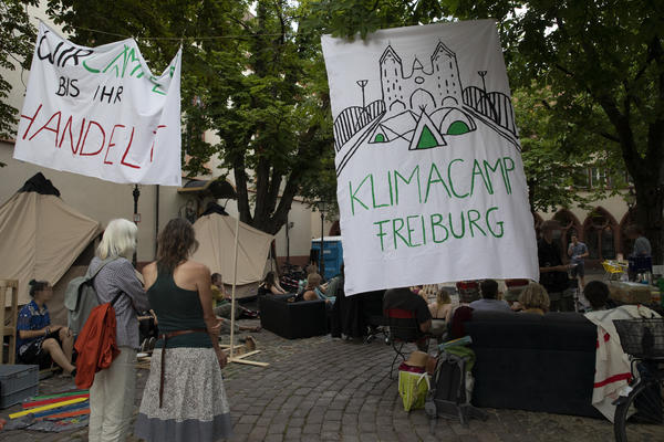 Es sind zwei Menschen vor an zwei auf einer Schnur befestigten Transparenten zu sehen. Auf dem linken Transpi steht &quot;Wir campen bis ihr handelt!&quot;, auf dem Anderen: &quot;Klimacamp Freiburg&quot; mit einer Sillhouette der Blauen Brücke und drei Zelten darauf.