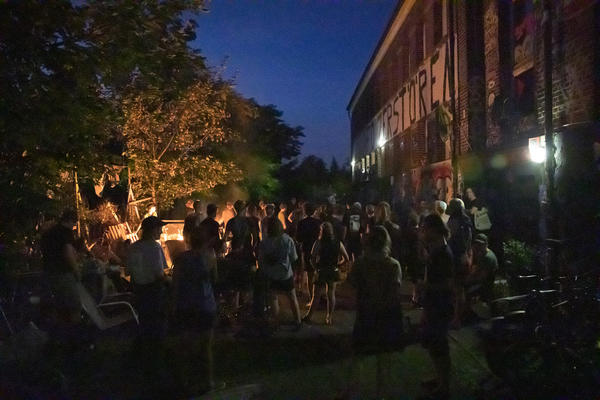 Blick in den Vorhof der KTS bei Nacht. Rechts das besprühte Specksteingebäude, links erhellt eine Feuertonne die Szene. Der Vorhof ist voll mit Menschen, die meisten stehen mit dem Rücken zur Kamera.