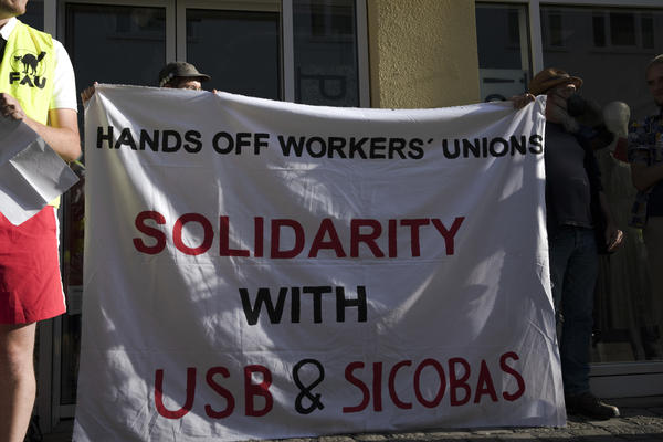 Zwei Personen, halten und befinden sich hinter einem weißen Transparent auf dem steht in schwarz und rot: Hands off workerss unions - Solidarity with USB &amp; SICOBAS. 
