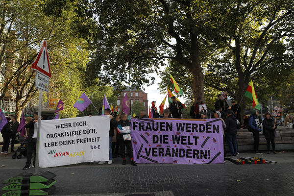 Demonstrant*innen am Platz der Alten Synagoge in Freiburg auf der Seite zur Bertoldstraße. Es sind kurdische Fahnen zu sehen und zwei Transparente. Auf dem rechten steht: &quot;Frauen werden die Welt verändern!&quot;, in Großbuchstaben, auf violettem Stoff.