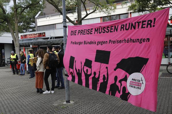 Rechts vorne ist ein rosa Transparent zu sehen mit &quot;Die Preise Müssen Runter! Freiburger Bündnis gegen Preiserhöhungen&quot; und unten Sillhouetten von Demonstrant*innen. Links, im Hintergrund sind Kundgebungs-Teilnehmende zu sehen.