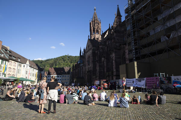 Blick auf dem Münsterplatz mit dem hinteren Teil des Münsters im Hintergrund. Rechts sind Transparente und eine kleine Bühne zu erkennen. Vorne sitzende und paar stehende Menschen, die einer redenden Person zuhören.