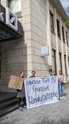 Menschen protestieren mit einem Banner mit der Aufschrift Deutsche Bank finanziert Klimakatastrophe und einem Schild mit der Aufschrift Pakistan is our story vor der Deutschen Bank Filiale in Freiburg.