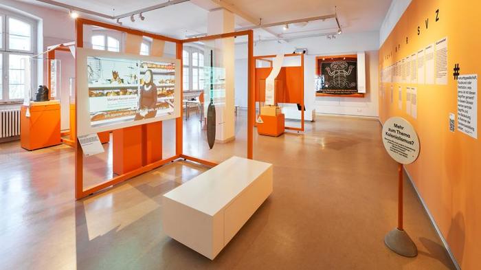  Blick in einen in Orange getauchten Ausstellungsraum gefüllt mit Vitrinen und Projektionsflächen, auf denen Filme laufen 
