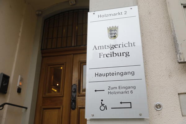Türschild des Amtsgerichts Freiburg, mit Wappen von Baden Württemberg. Links daneben und dahinter die schwere Holztür mit kleinen Fenstern darin. Eine Türklingel ist auch zu sehne.