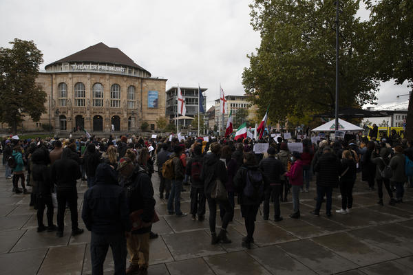 Es ist die Menschenmenge auf dem Platz der Alten Synagoge zu sehen. Mehrere tragen iranische Flaggen (ohne Hohheitsabzeichen), viele halten weiße Schilder in die Höhe. Links im Hintergrund ist das Freiburger Stadttheater zu sehen. 