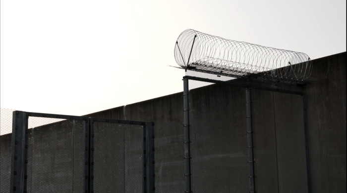 Hohes Gittertor, rechts etwas höher mit spiralförmigem Stacheldraht. Dahiner Gefängnismauer.