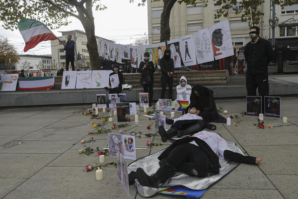 Mehre Menschen mit übergezogenen weißen und blutverschmierten Hemden liegen auf dem Boden. Kerzen, Schilder mit Gesichtern und namen sind verteilt. Rechts steht eine Person mit weiß bemaltem Gesicht und schwarzer Kleidung an einem Strick.