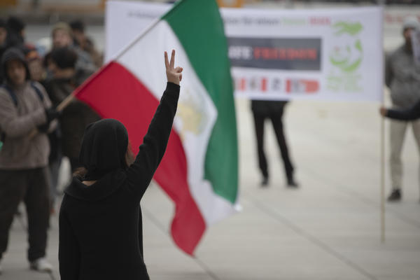 Eine Person mit langen braunen haaren, schwarzer Filzmütze und schwarzer Kleidung ist von hinten zu sehen, wie sie ihre rechte Hand zum Victory &amp; Peace - Zeichen in die Höhe streckt. Im Hintergrund unscharf eine Iran-Fahne zu sehen und weitere Menschen.