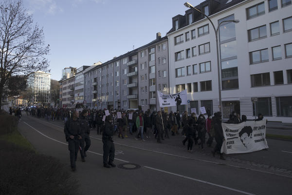 Demonstrationszug in der Friedrichstraße in einer schrägen Seitenansicht. Weißes Fronttransparent weist auf den Mord an Oury Jalloh hin und ein ebenfalls weißes Hochtransparent auf Polizeigewalt insgesamt.