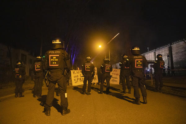 Polizei mit Helmen blockiert Demonstration, Banner mit &quot;Mauern und Grenzen sprengen&quot;, eine Sylvesterrakete fliegt über die Mauer der JVA Freiburg