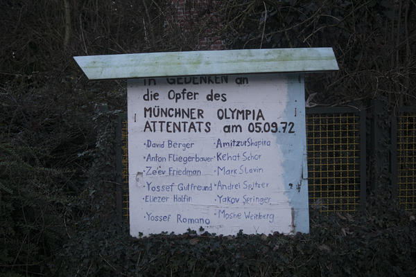 Ein überdachtes Schild in Lützerath gedenkt an die Opfer des Münchner Olympia Attentats am 05.09.1972. Es sind 12 Namen zu lesen. Im Hintergrund sind Büsche und ein gelber Metallzaun zu sehen.