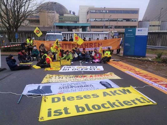 Menschen sitzen und stehen mit Bannern, Fahnen und Schildern vor dem Eingangstor des Atomkraftwerks Emsland.
