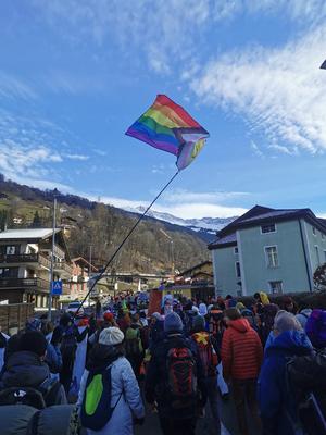 Menschen wandern als große Gruppe nach Davos, im Hintergrund Schnee bedeckte Berge, eine LGBTQIA* Flagge wird geschwenkt.