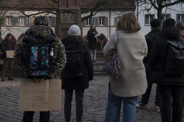 Vier Menschen stehen mit dem Rücken zur Kamera im Bild. Person links trägt Leojacke und ein Pappschild vor dem Gesäß: 10 Jahre ich hier immer noch keine Papiere. Im Hintergrund Rednerin vor dem Brunnen auf dem Rathausplatz.