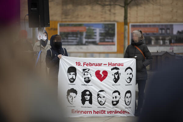 Zwei Personen halten ein Transparent mit neun aufgemalten Portraits der Getöteten. Oben in rot mit einem zerbrochenem Herzen &quot;19. Februar - Hanau&quot;, unten: &quot;Erinnern heißt verändern.&quot;