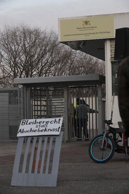 Das Eingangstor der LEA Freiburg im Hochformat. Vorne ein Schild mit Bleiberecht statt Abschiebeknast. Hinten das offizielle Schild und ein*e Bewohner*in, die gerade von Security durchsucht wird.