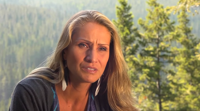 Still from the documentary PLACEnta by Jules Koostachin (2012): The filmmaker talking to an interviewer in front of a forest. She is looking worried and frowns.