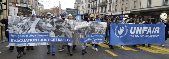 Demonstrierende auf der Straße mit blauem Banner: UNFAIR - The UN Refusal Agency